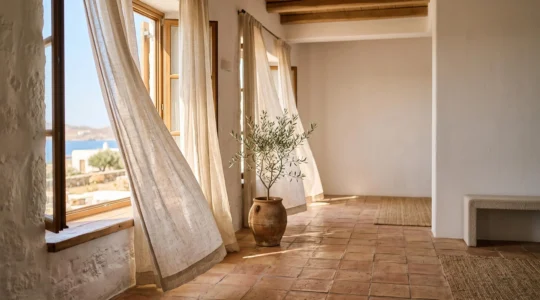 Salón mediterráneo luminoso con cortinas de lino natural ondeando junto a una ventana abierta en verano