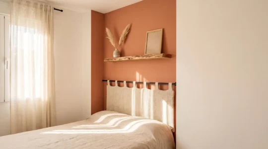 Rincón de dormitorio renovado con estantería flotante de madera clara, cabecero textil y luz natural cálida, transformado con un presupuesto reducido.